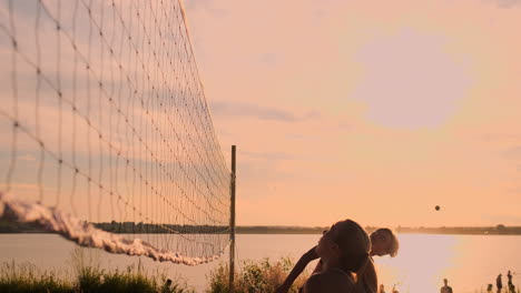 SLOW-MOTION-LOW-ANGLE-CLOSE-UP-LENS-FLARE:-Young-female-volleyball-players-pass-and-spike-the-ball-over-the-net-on-a-sunny-summer-evening.-Fit-Caucasian-girls-playing-beach-volleyball-at-sunset.